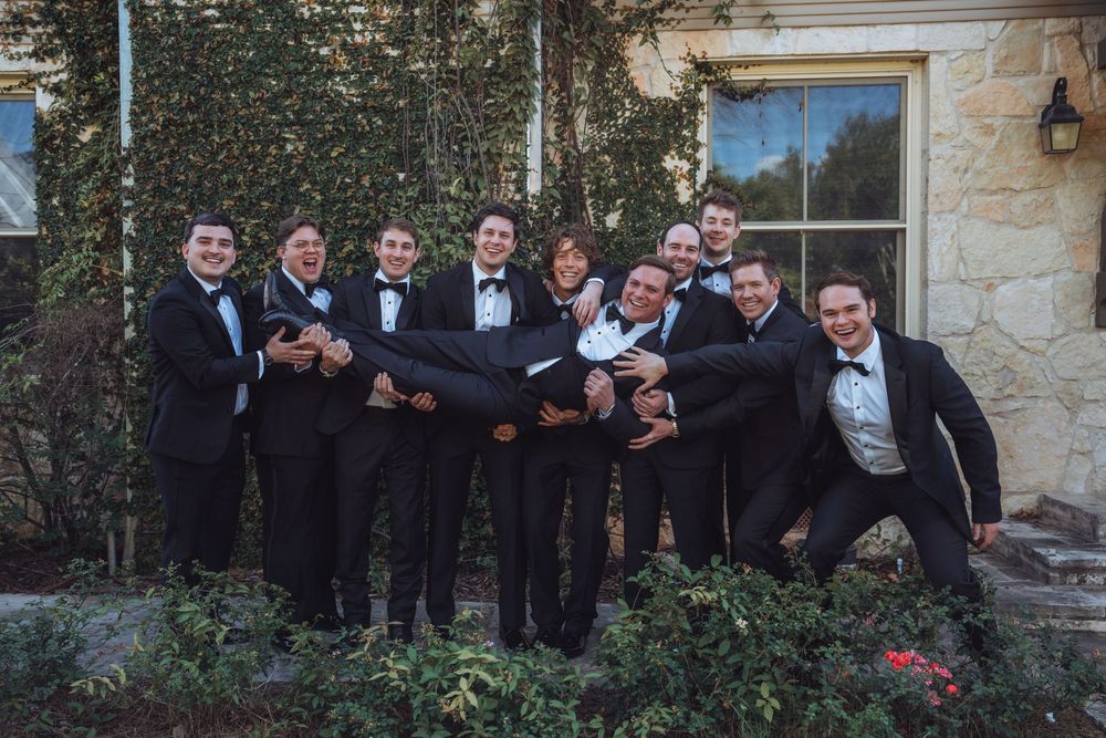 Groomsmen in black tuxedos lifting groom outside ivy-covered venue in fun wedding photo.
