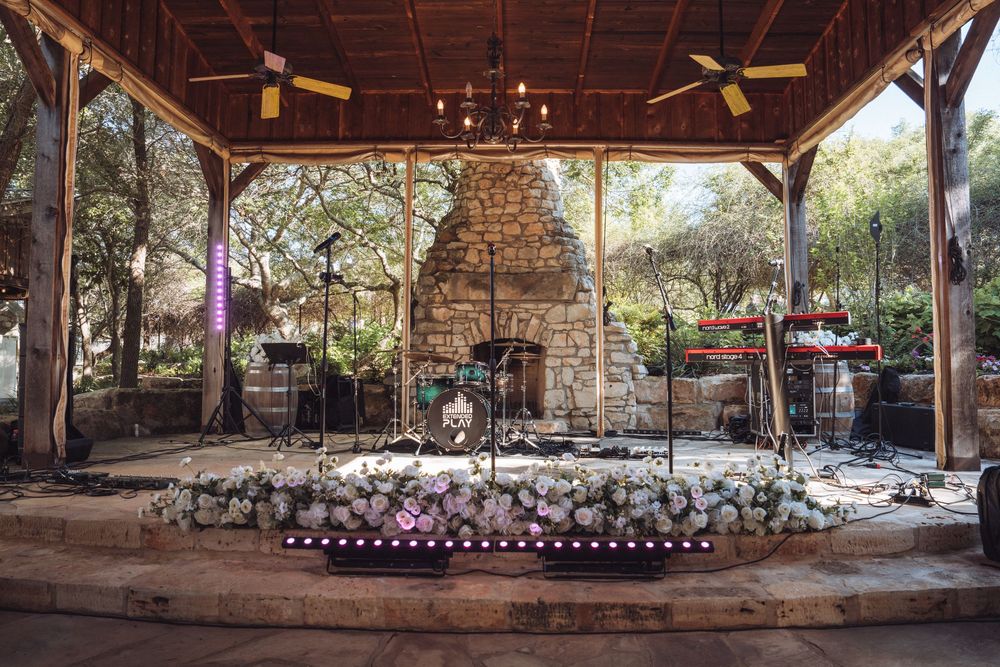 Rustic outdoor wedding stage with stone fireplace, floral decorations, and band setup under a wooden pavilion.