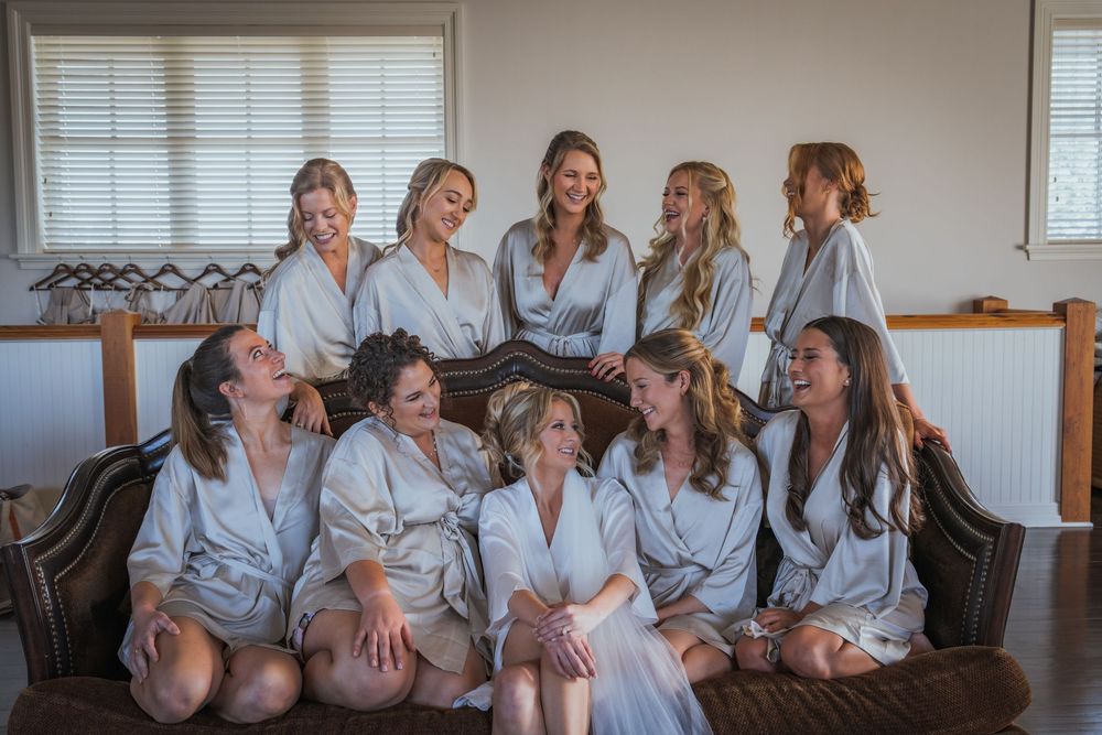 Bridesmaids in matching satin robes laughing together on a couch during wedding preparations.