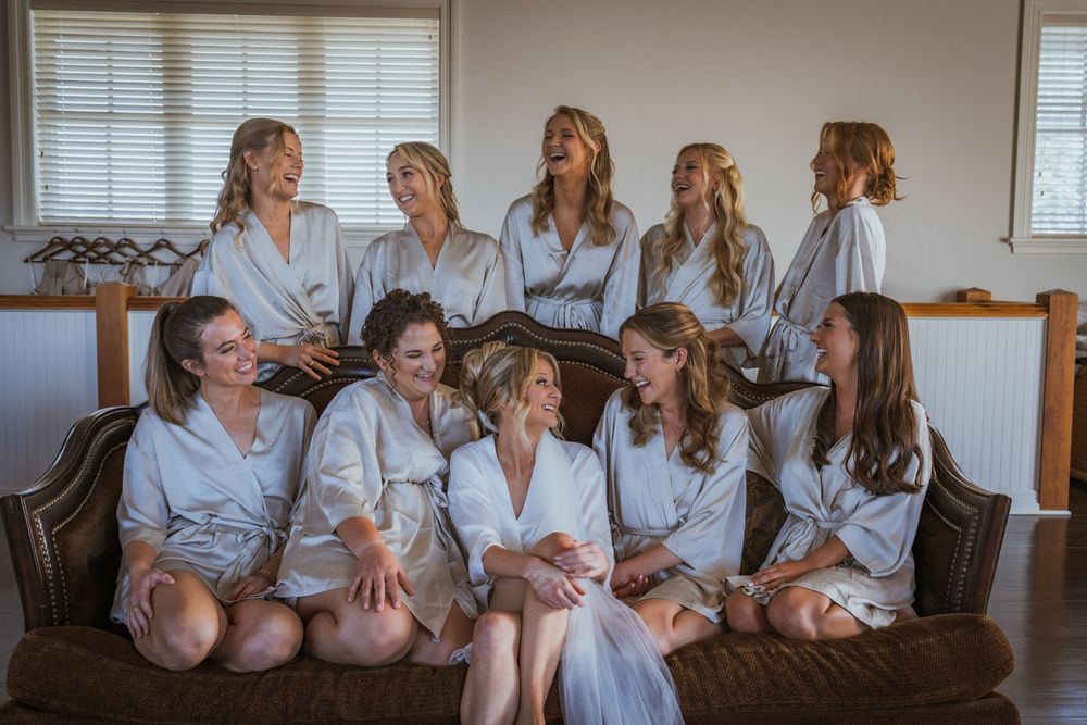 Bridesmaids in matching white robes laughing together on a couch during wedding preparations indoors.