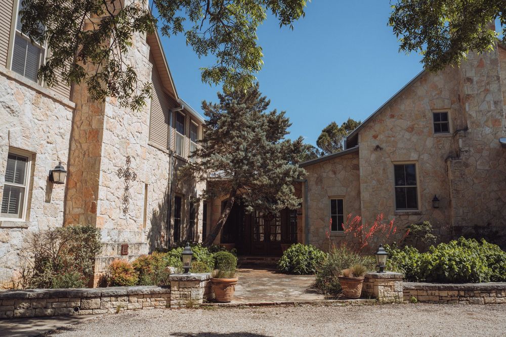 Rustic stone building courtyard with lush trees, potted plants, and warm natural light on a clear sunny day.