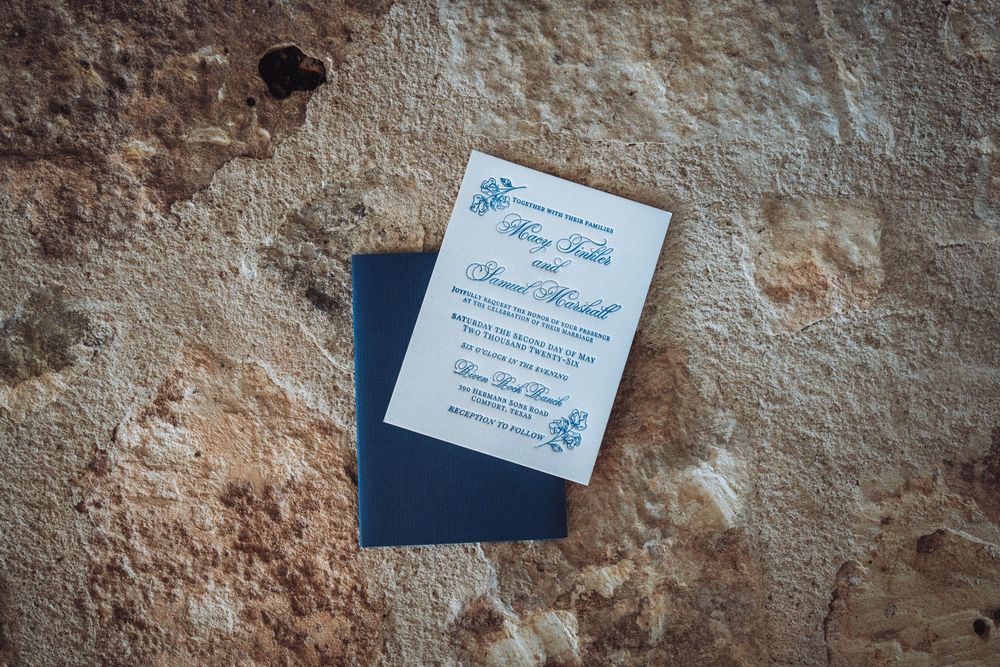 Elegant wedding invitation with blue floral details resting on a rustic stone surface with a navy envelope.