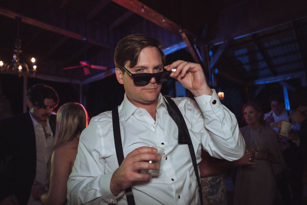 Man in white shirt and suspenders adjusts sunglasses while dancing at a wedding reception.