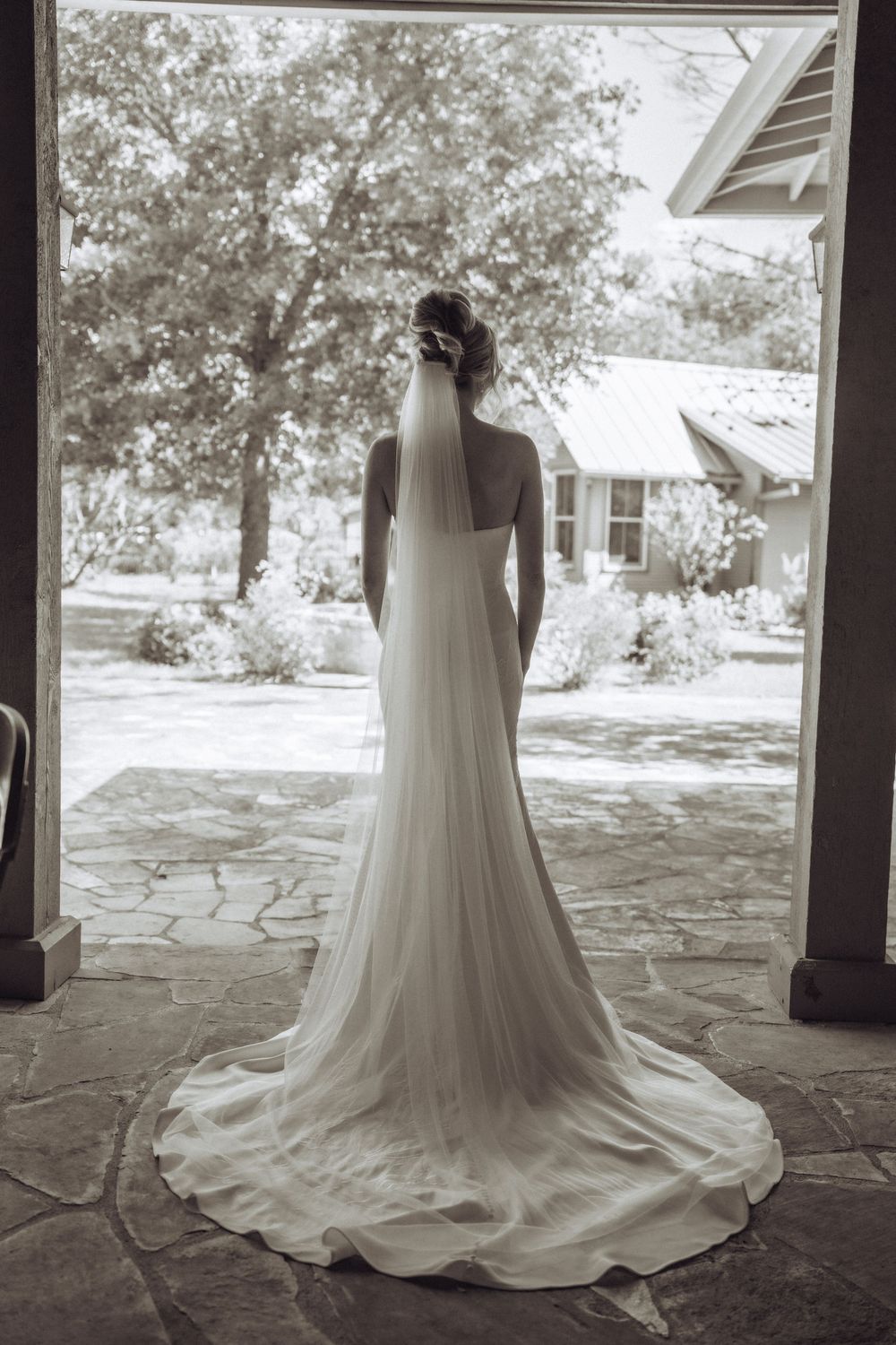 Elegant bride in a flowing white wedding gown and veil stands in a doorway overlooking a lush outdoor venue.