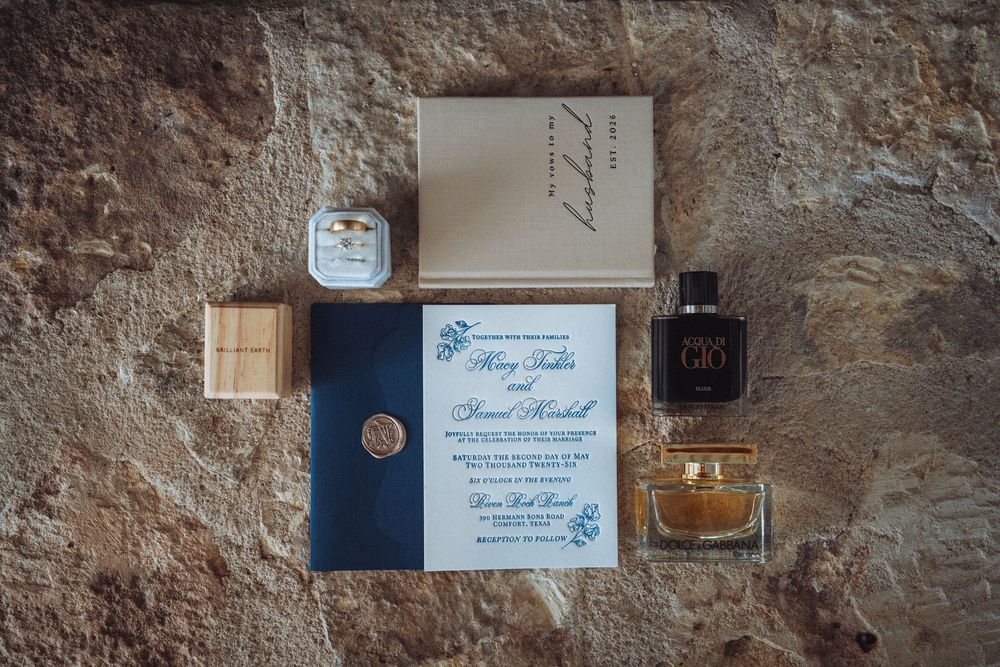 Wedding flat lay featuring blue invitation, cologne, ring box, and accessories arranged on stone surface.