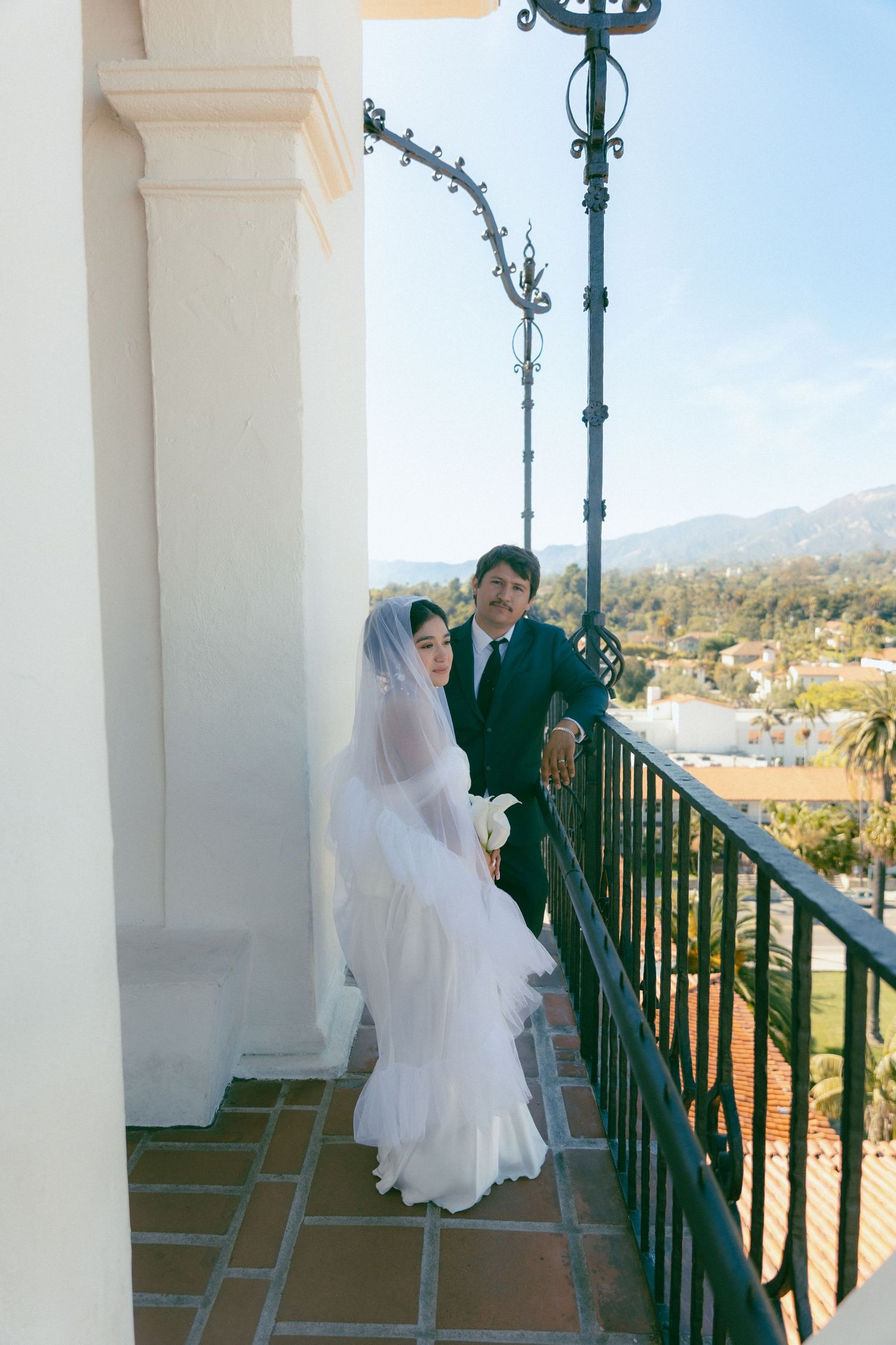 Bride and groom pose on a sunlit balcony with mountain views, bride in white gown and veil holding bouquet.