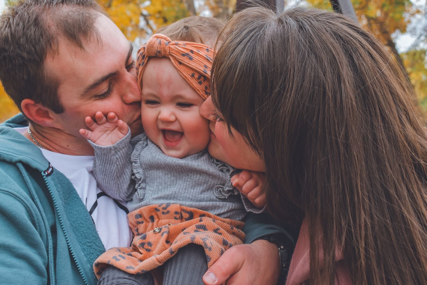 Happy baby laughing as parents kiss her cheeks outdoors in autumn park with golden foliage background.