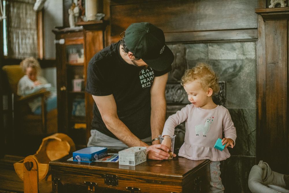 A father helps his toddler at a wooden table in a rustic room, with a woman visible in the background. documentary in home photography