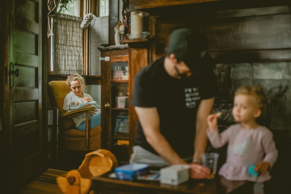 A bearded man and toddler girl stand in a rustic cabin while an mother tends a baby in a chair in the background. documentary in home photos
