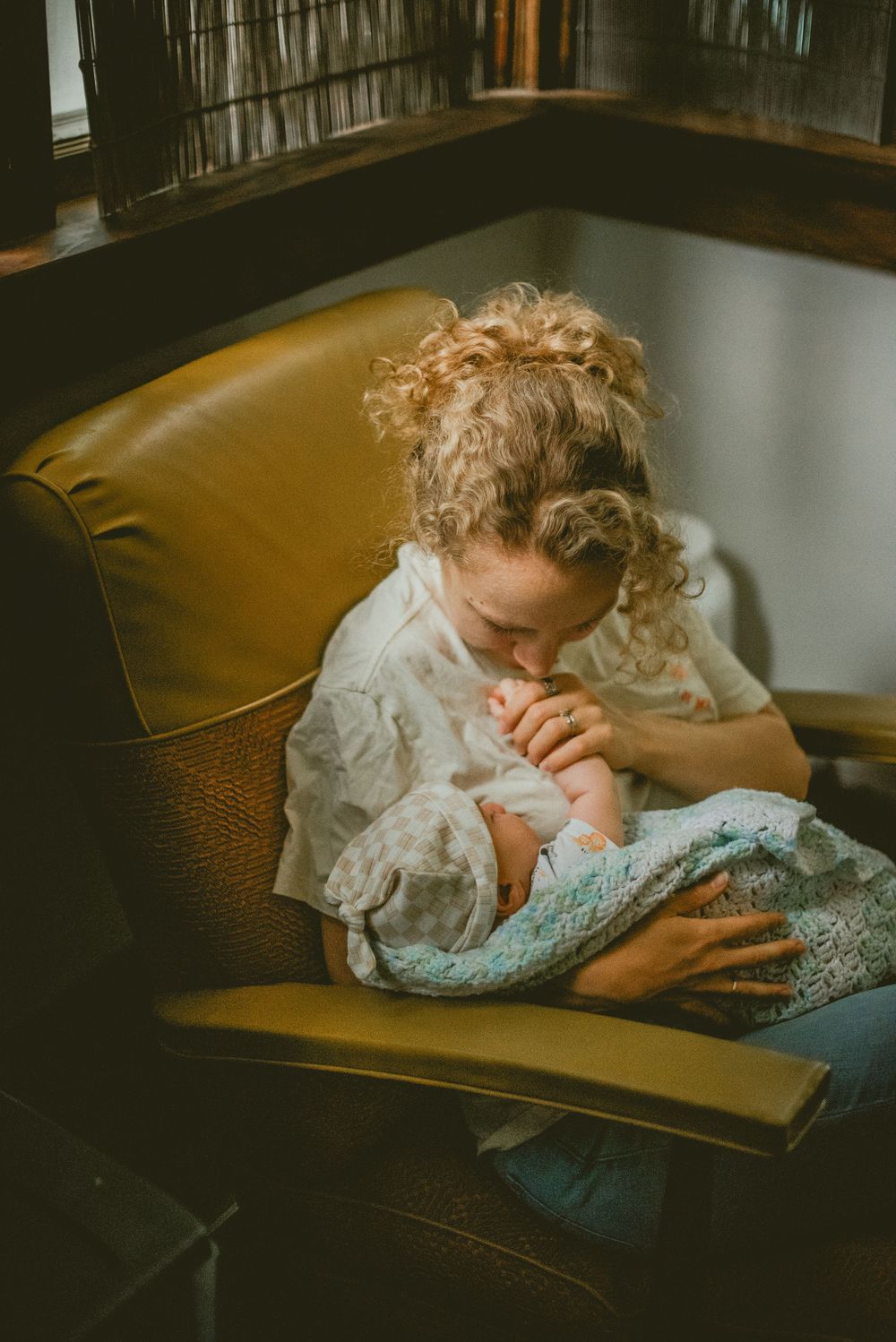 Mother with curly hair sitting in a yellow armchair, gently kissing a newborn baby wrapped in a blue blanket. documentary in home photos