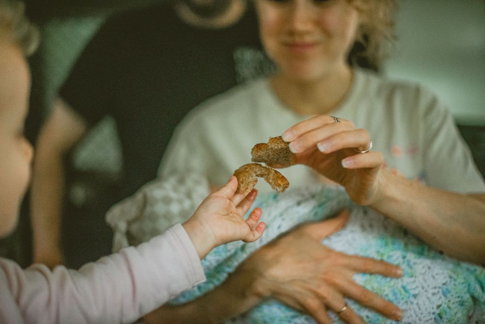 A family shares a cookie with a young child, with a smiling woman holding a baby in the background. documentary in home photography