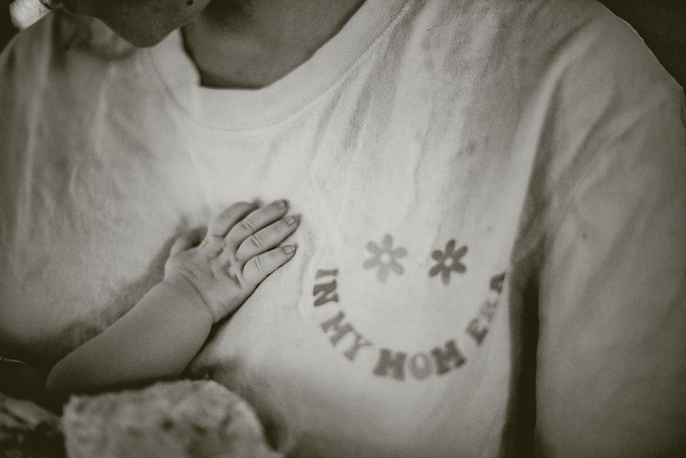 Black and white photo of a newborn baby's hand resting on a mother's chest wearing an 'In My Mom Era' t-shirt. documentary in home photos