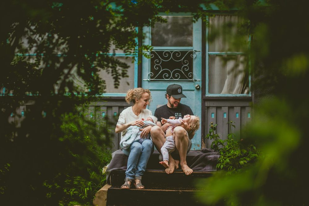 Family sitting on front porch steps with two young children, surrounded by lush green trees near a blue door documentary in home photography