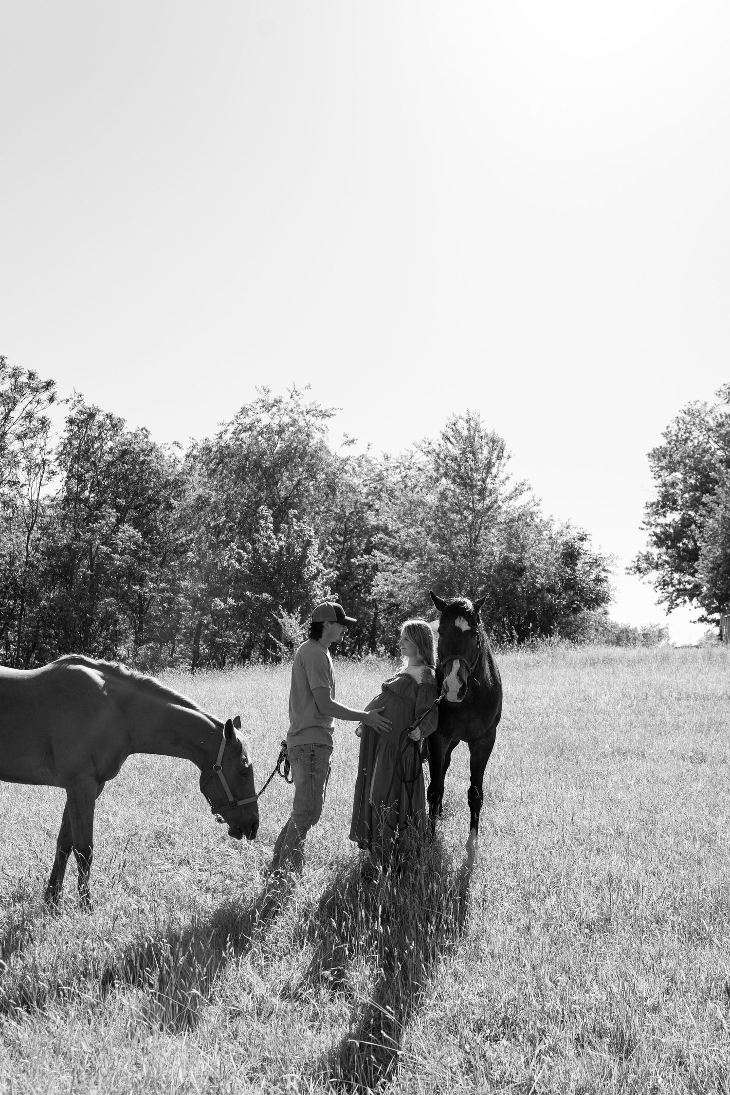 Black and white photo of a couple with two horses in a grassy field surrounded by trees.