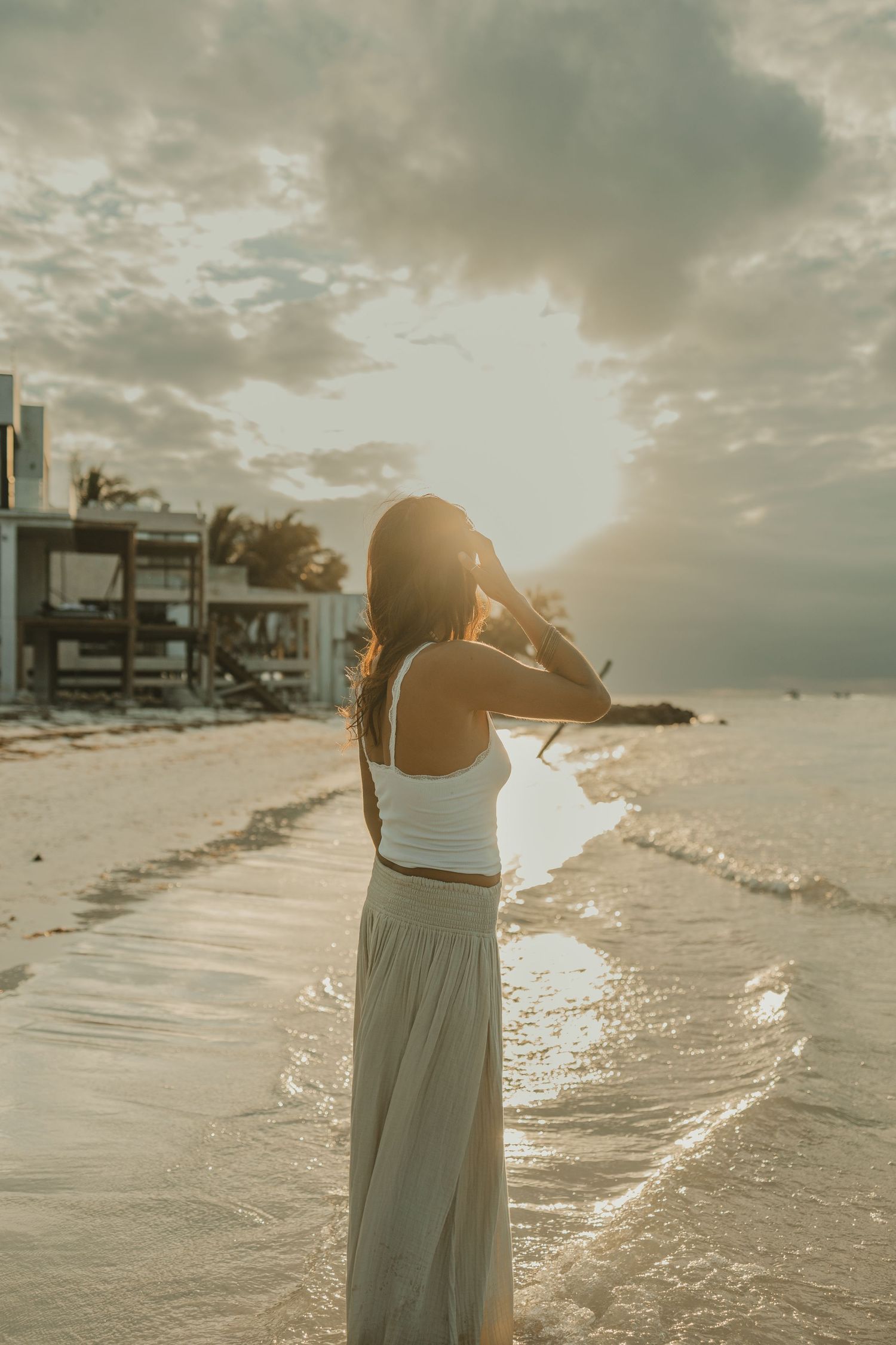 Woman in white crop top and maxi skirt standing on a beach during a golden sunset.