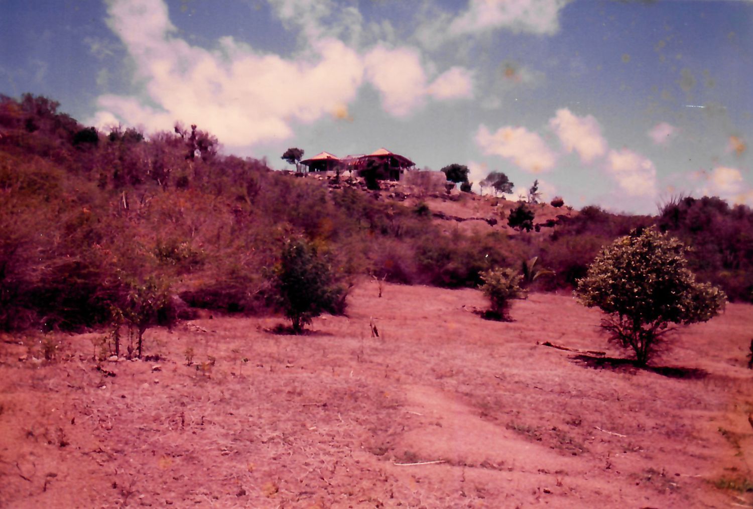 Arid hillside landscape with sparse trees, red soil, and a small structure on the hilltop under a partly cloudy sky.