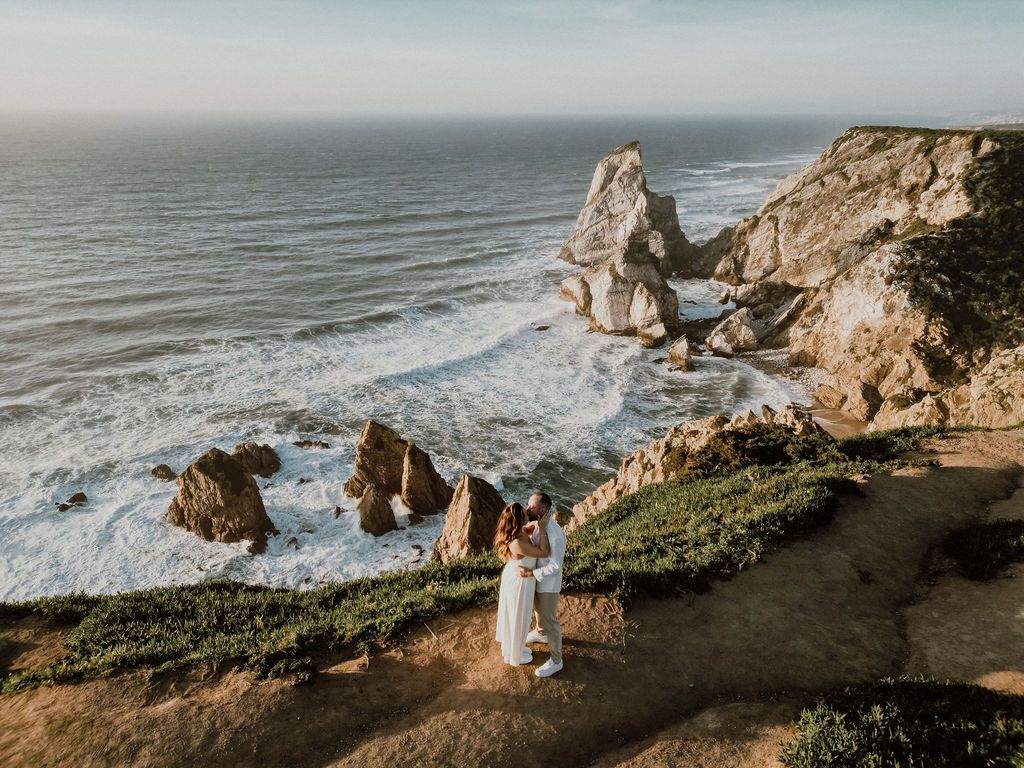 Couple standing on a dramatic coastal cliff overlooking rocky sea stacks and ocean waves in Portugal.