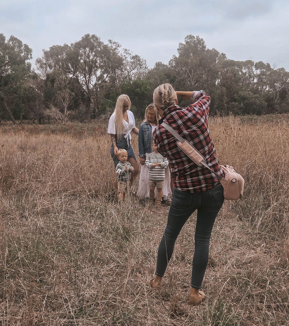 A photographer captures a family walking through a golden field with trees in the background on a cloudy day.