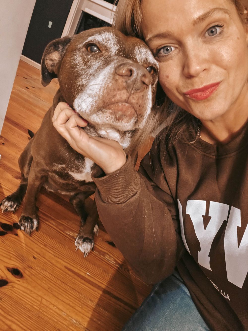 Woman in NYC sweatshirt smiling with her senior brown pitbull dog on hardwood floors.
