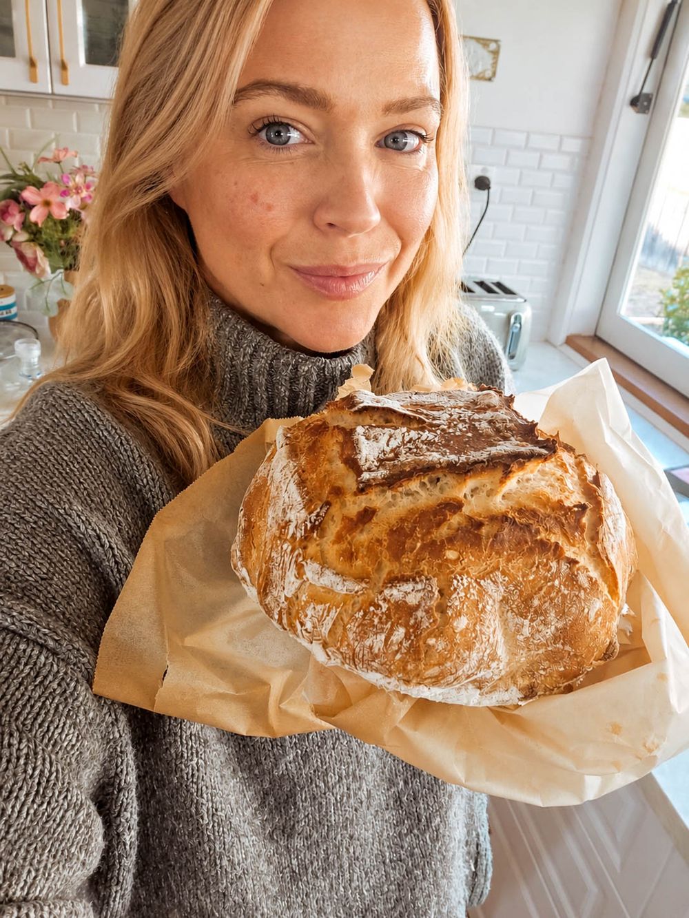 Woman in gray sweater proudly holding a freshly baked rustic sourdough loaf on parchment paper in a bright kitchen.