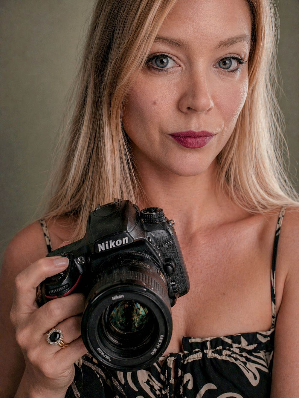 Blonde woman with blue eyes holding a Nikon DSLR camera, wearing a floral top and dark lipstick.
