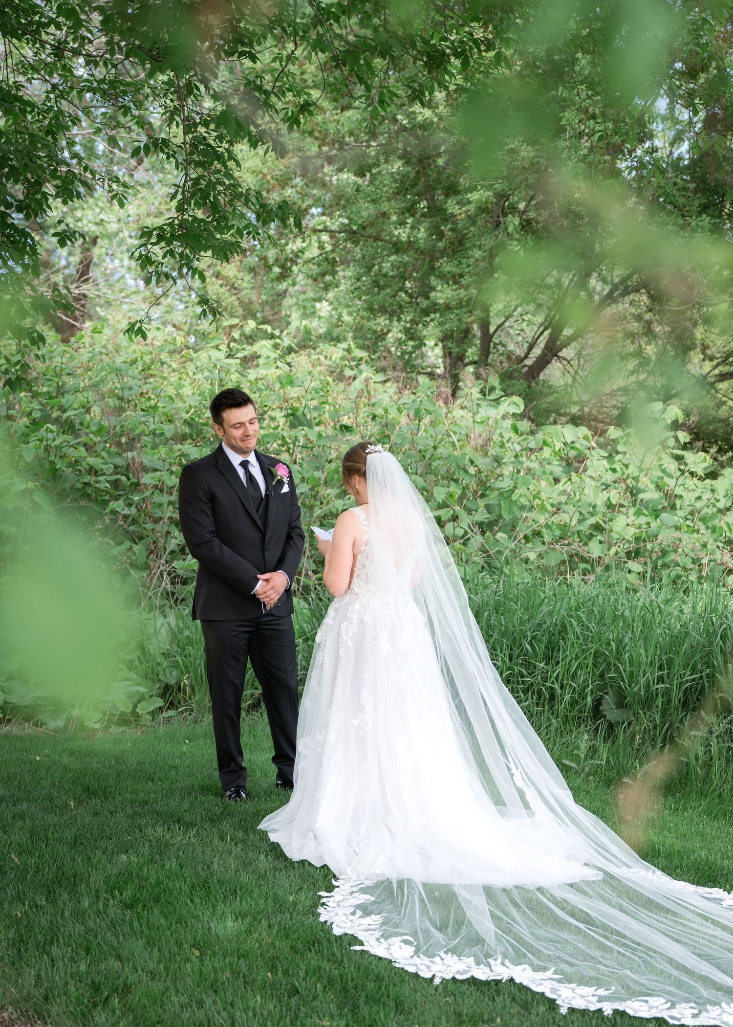 Bride in white gown with long veil stands with groom in black suit outdoors amid lush green trees.