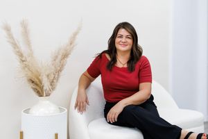 Woman in red top sitting relaxed on white chair beside pampas grass decor in bright modern studio.