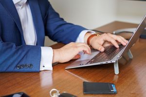 A businessman in a blue suit typing on a laptop computer elevated on a stand at a wooden desk.