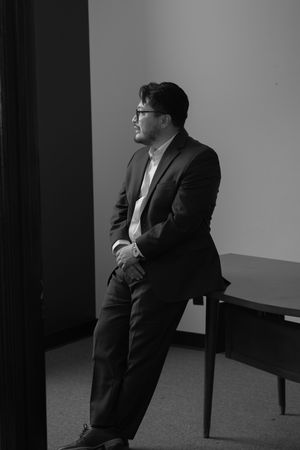 Man in suit and glasses sitting pensively in black and white photo in a minimalist office setting.