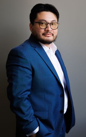 Professional man in blue suit and glasses poses confidently against gray background for business portrait.