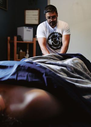 A massage therapist in a white graphic tee performs a therapeutic massage on a client covered with a blanket.