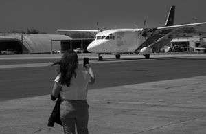 Black and white photo of a woman photographing a small propeller aircraft on a tarmac.