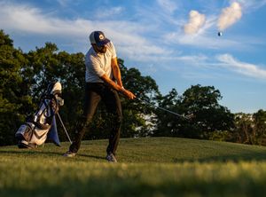 Golfer hitting an iron shot on a golf course at dusk with bag nearby.
