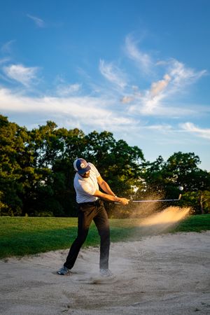 Golfer hitting a bunker shot with sand exploding under blue sky.