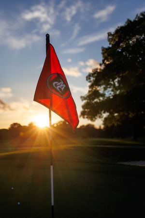 Red golf flag with CGA logo backlit by golden sunset on lush green course with trees silhouetted against blue sky.