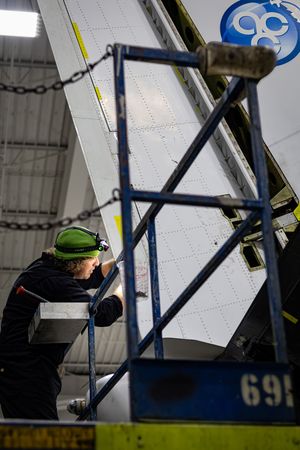 Aircraft mechanic in green hard hat inspecting airplane wing in a maintenance hangar.
