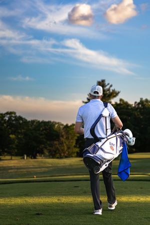 A golfer carries a white golf bag across a lush green fairway under a blue sky.