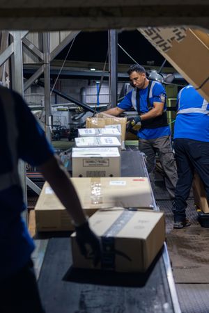 Warehouse workers in blue vests sort and scan packages on a conveyor belt.