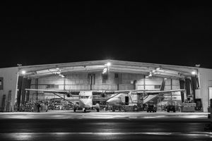 Black and white night shot of a twin-engine aircraft inside an illuminated airport hangar.
