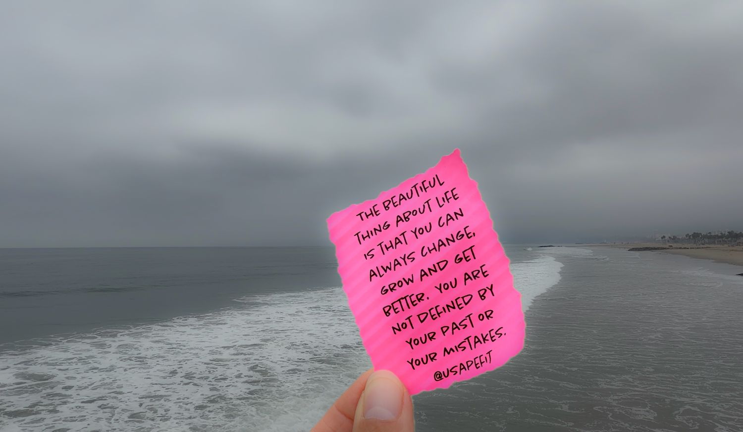 Hand holding a pink motivational note card against a cloudy ocean beach backdrop.