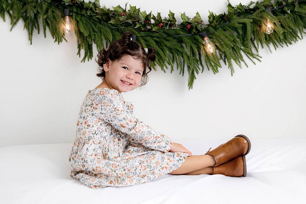 Young girl in floral dress and brown boots sitting under Christmas garland with fairy lights.