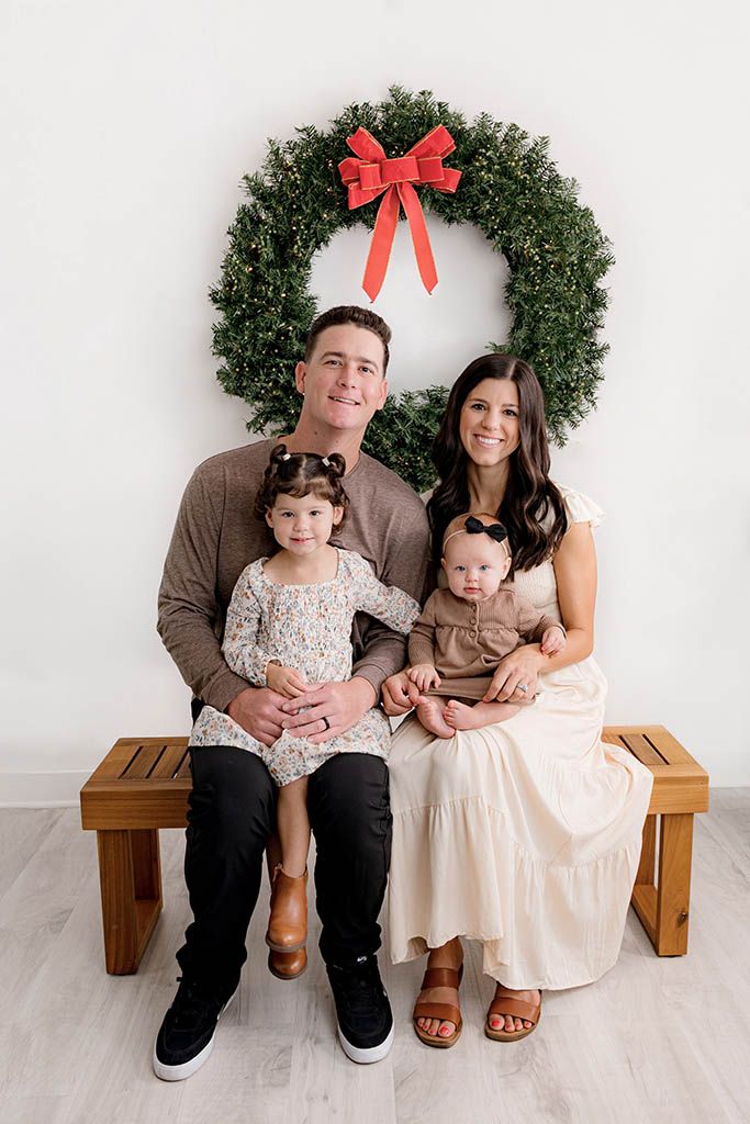 Family of four posing for a Christmas portrait in front of a large green wreath with a red bow.