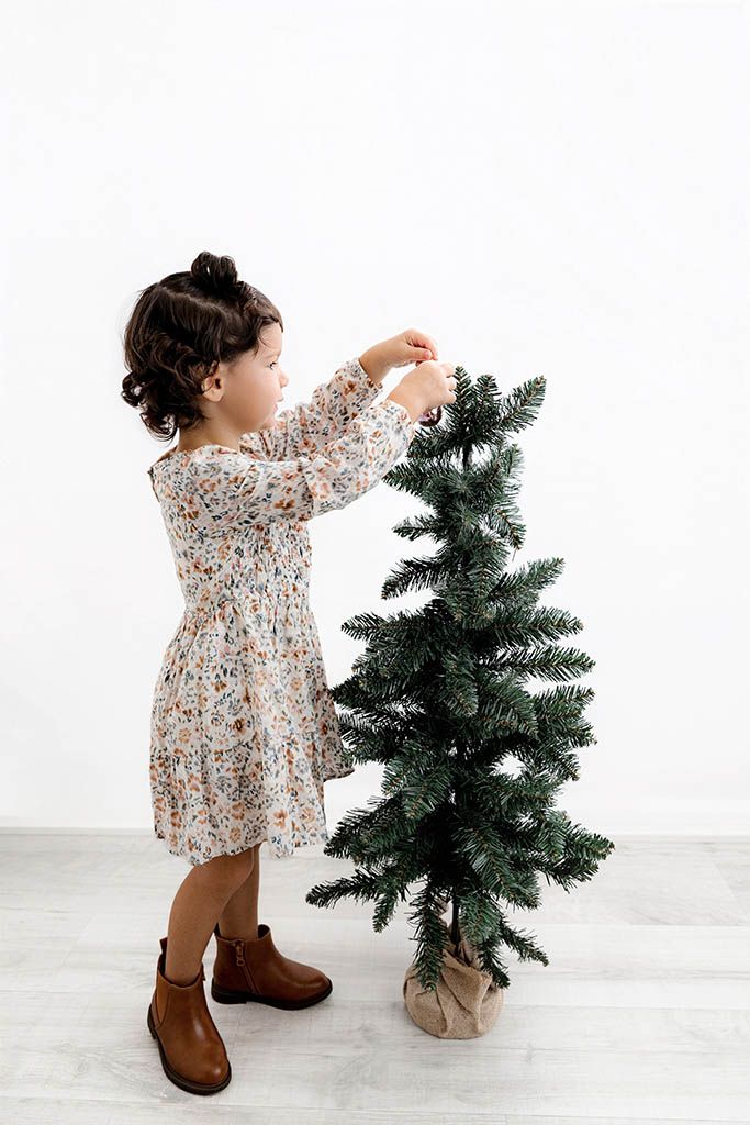 A toddler girl in a floral dress decorates a small Christmas tree against a white wall.