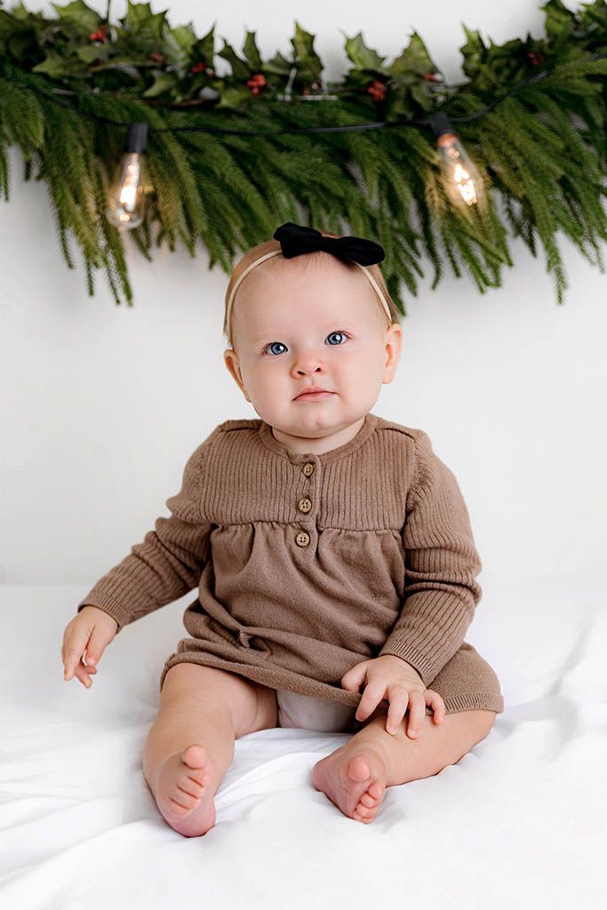 Baby girl in brown knit romper with black bow headband sits on white surface before Christmas garland.