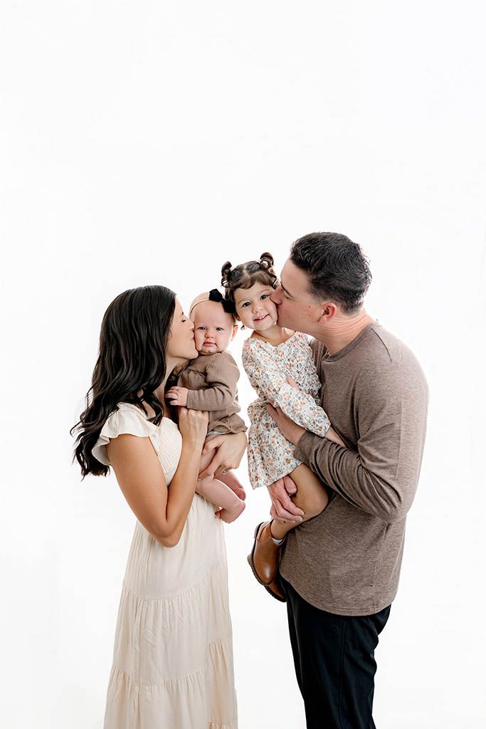 A happy family of four with parents kissing their two young children during a professional white studio photo session.