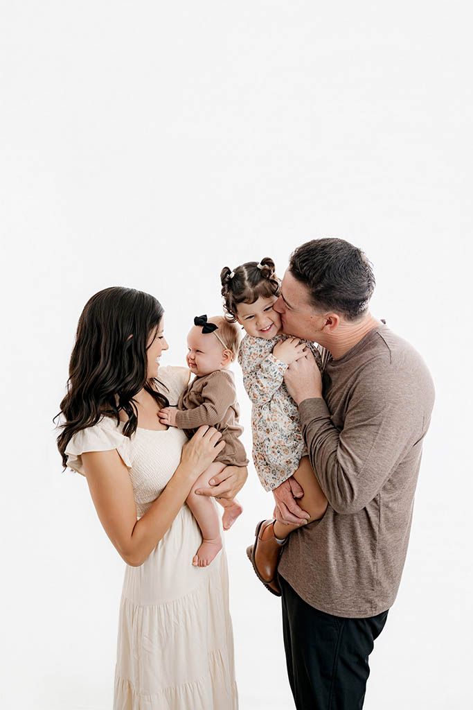 A happy family of four poses together, parents holding two young children in a bright white studio setting.