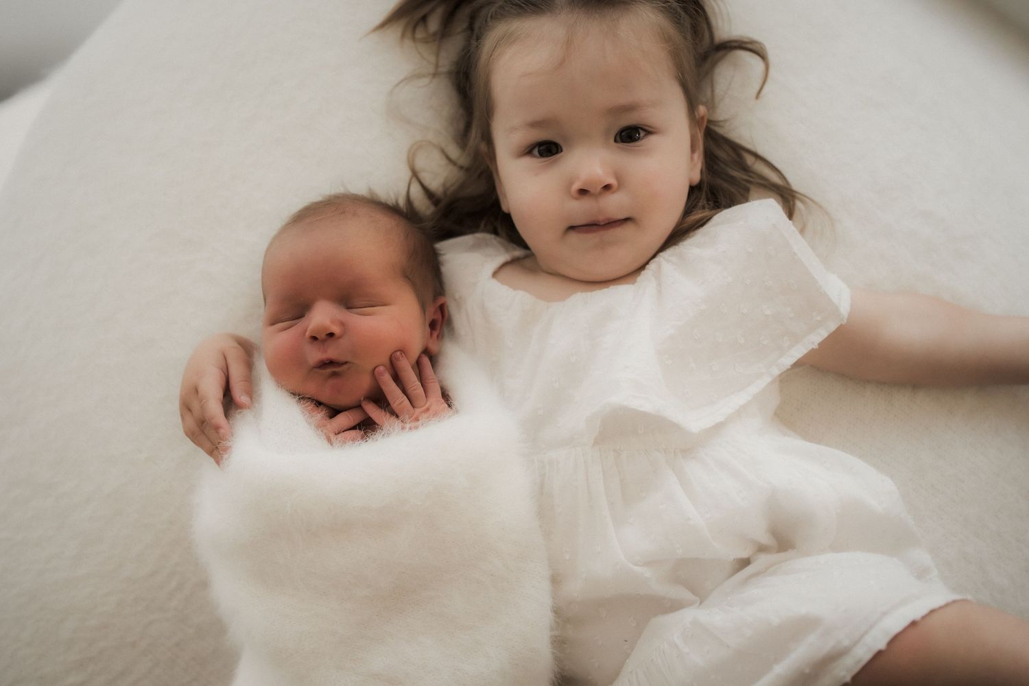 A toddler girl in a white dress lies next to a sleeping newborn wrapped in a white swaddle on a soft white surface in Buxton, Victoria
