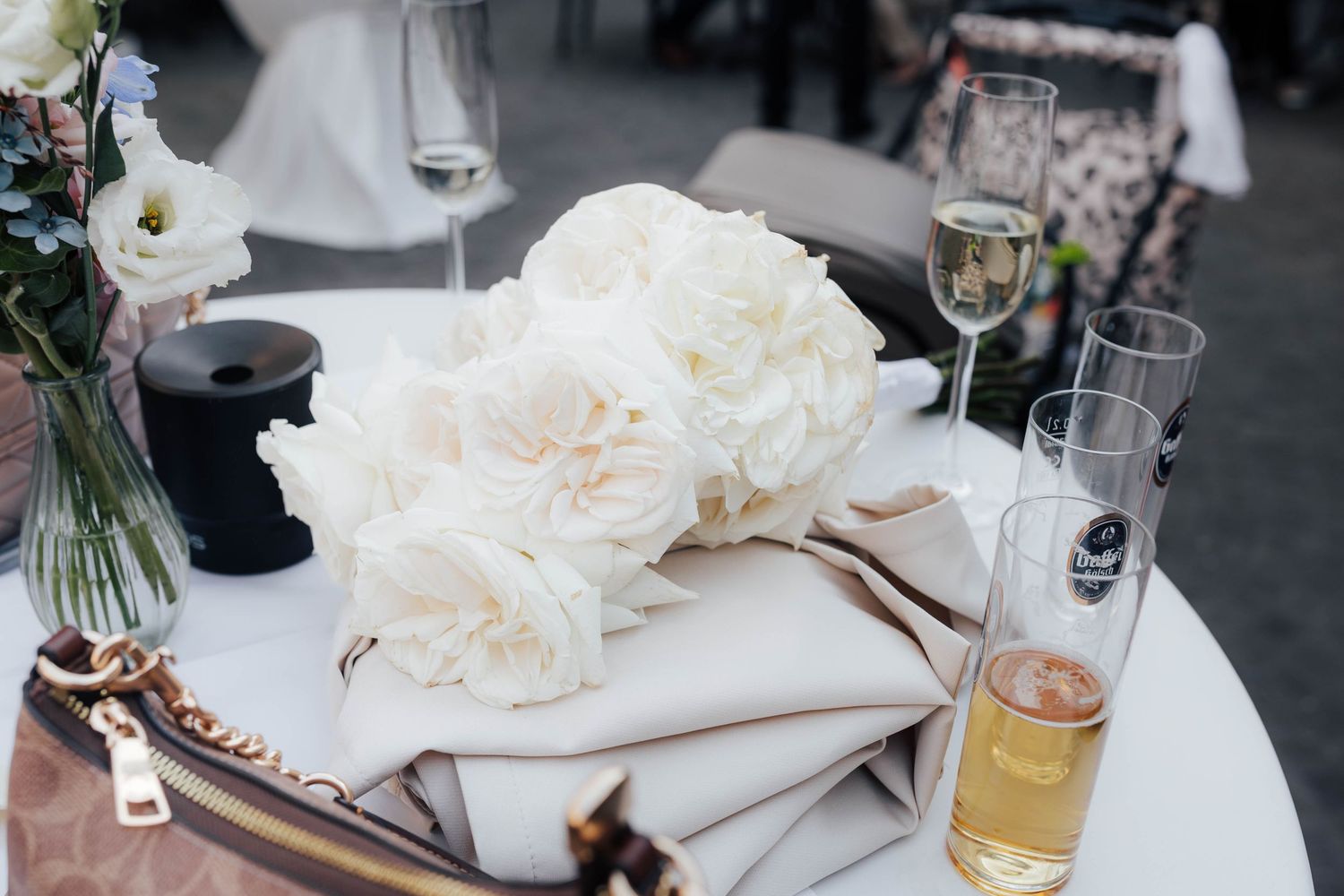 White peony bouquet resting on a beige purse beside champagne glasses on a white cocktail table at a wedding reception.