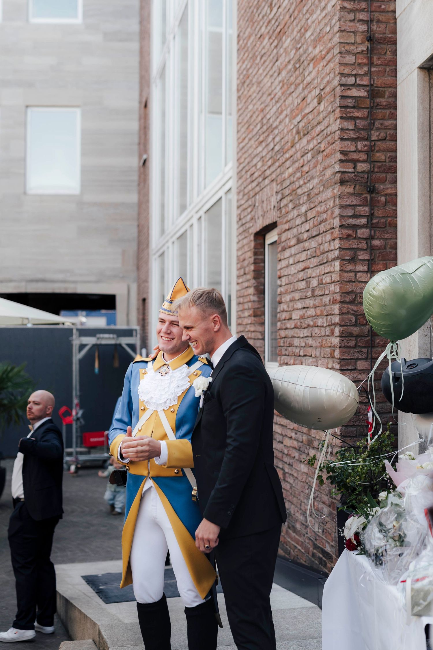 Two men in formal attire, one in a military-style uniform, stand together outside a brick building with balloons nearby.