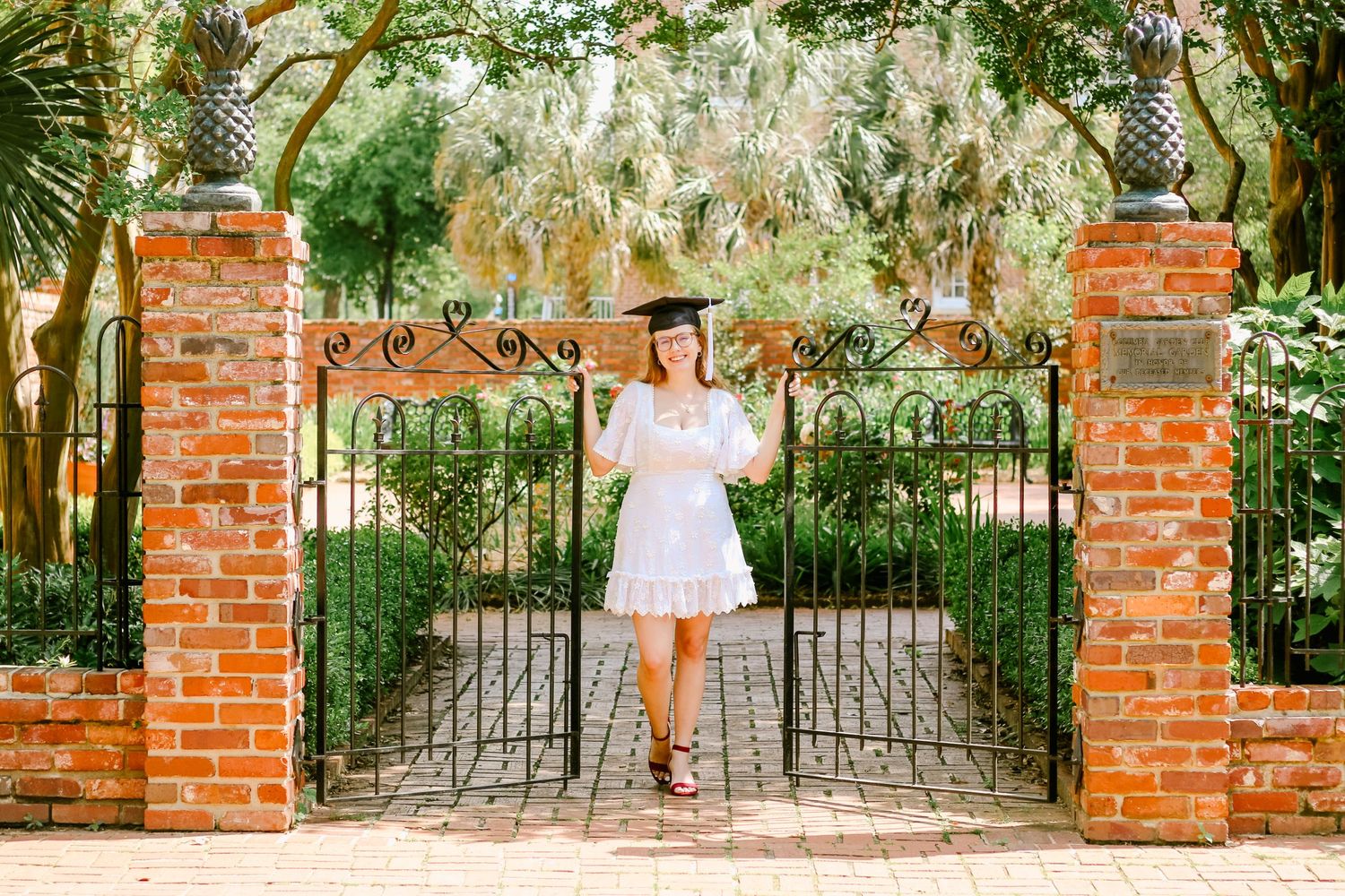 A person in white dress poses at ornate brick entrance gates with spanish moss overhead.